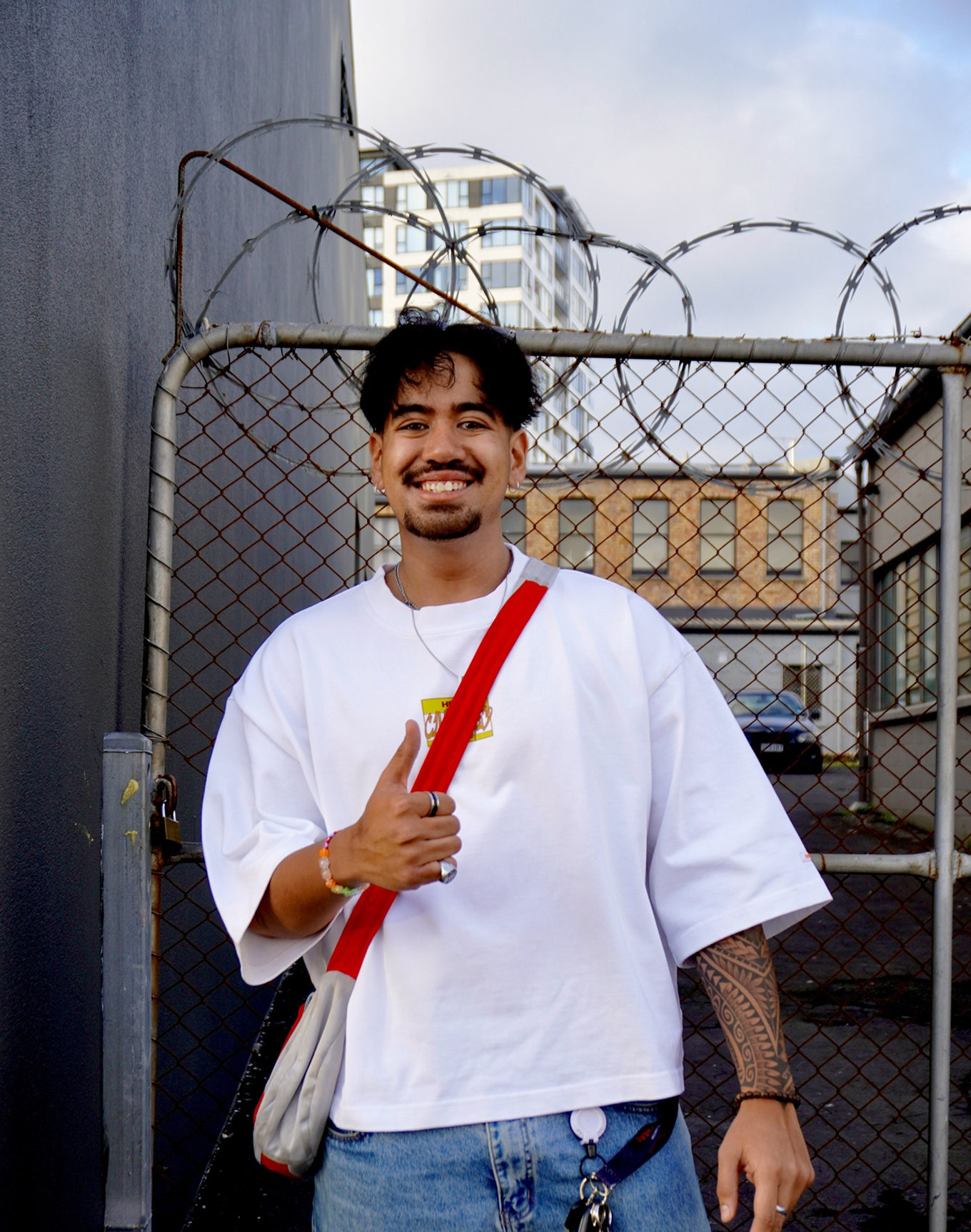 A young man white a red bag posing next to a fence. 