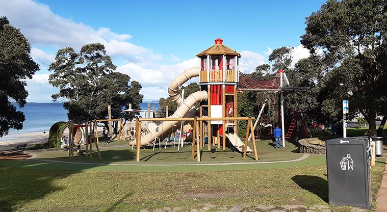 Picture of tower with several slides coming off it with swings in the foreground and Takapuna beach in the background