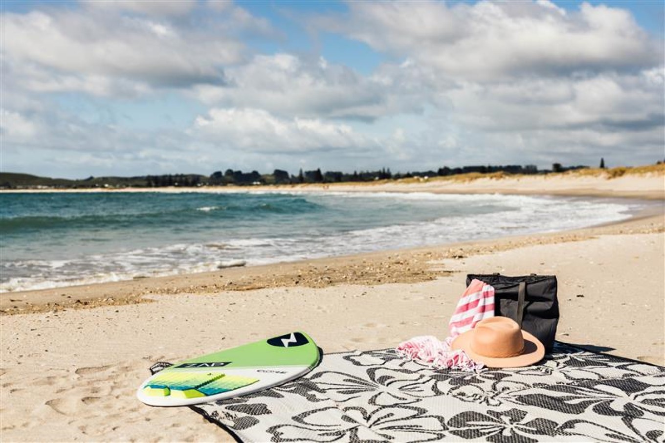 Green skim board, hat and bag on a floral picnic blanket at Omaha Beach, with the sea in the background.