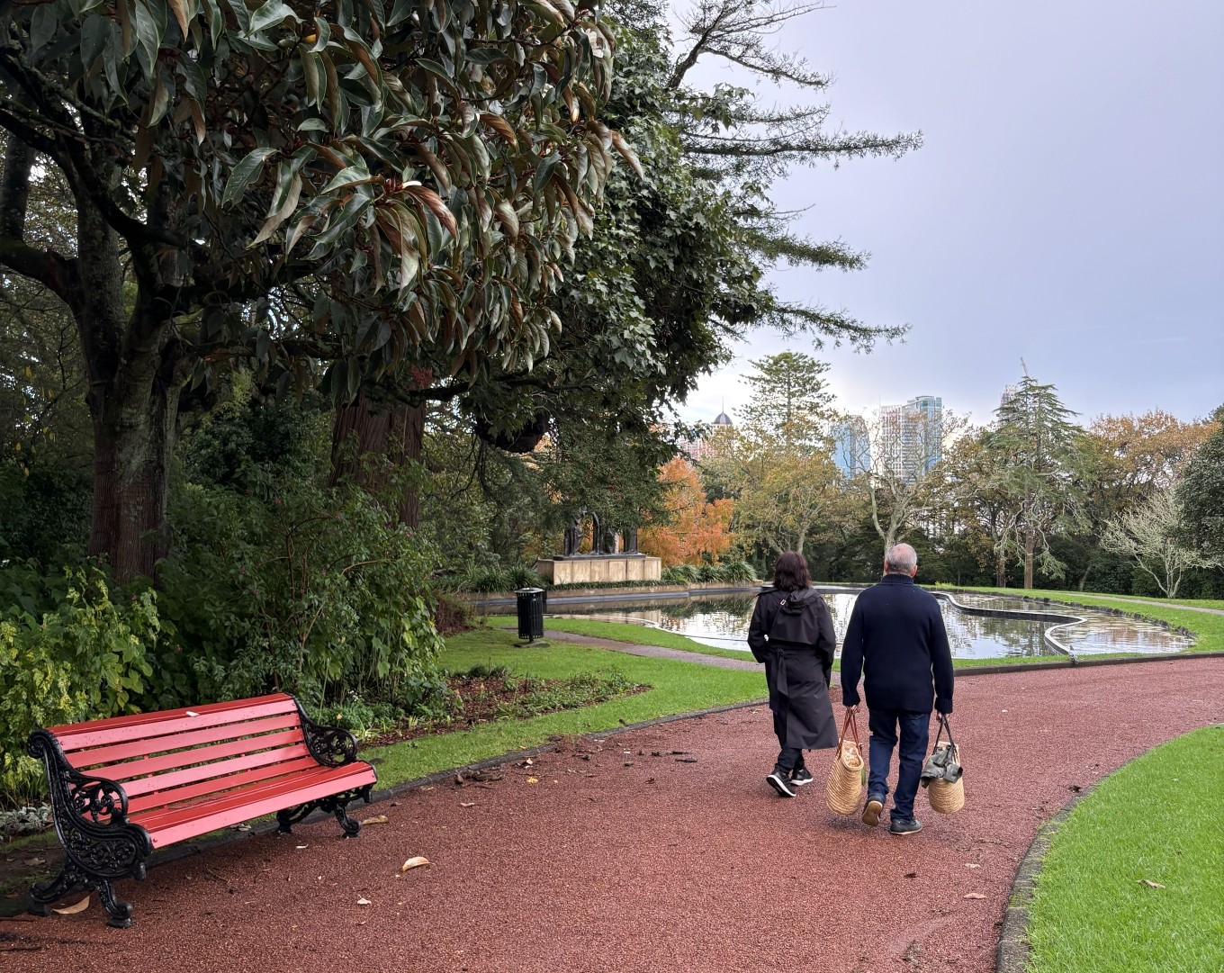 Two people walk along a path next to a park bench and pond.