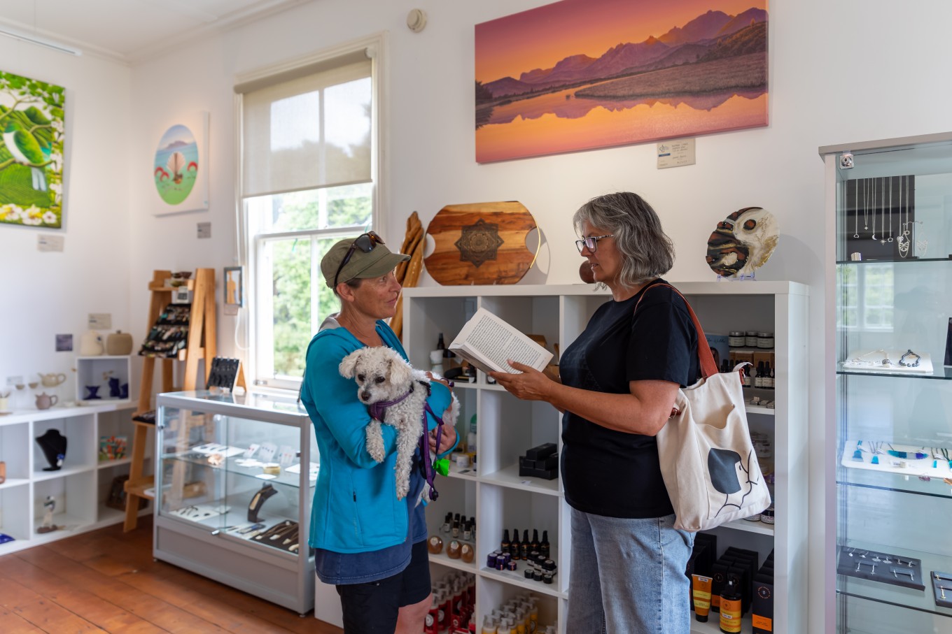 Two women talking in a gallery gift shop, one holding a dog and the other holding a book