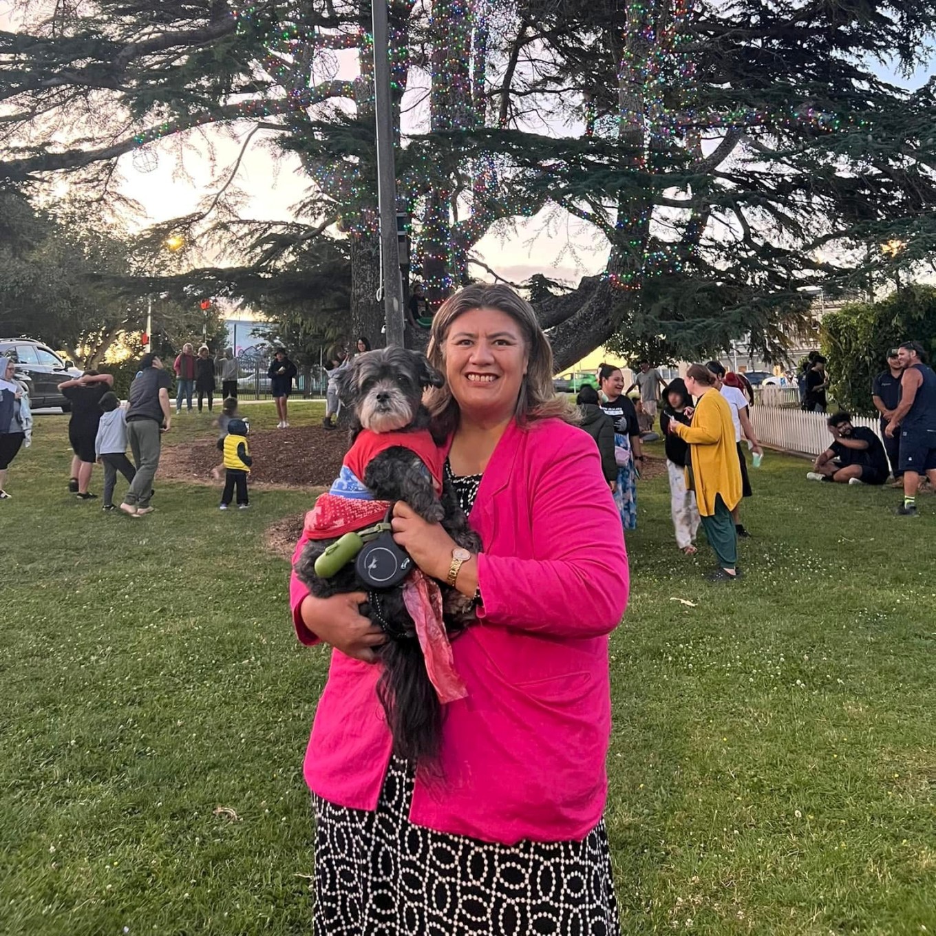 A lady in a pink shirt holding a dog at a park.